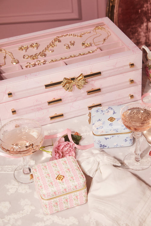 Pink jewelry box with gold accents on a table with glasses of champagne and flowers.